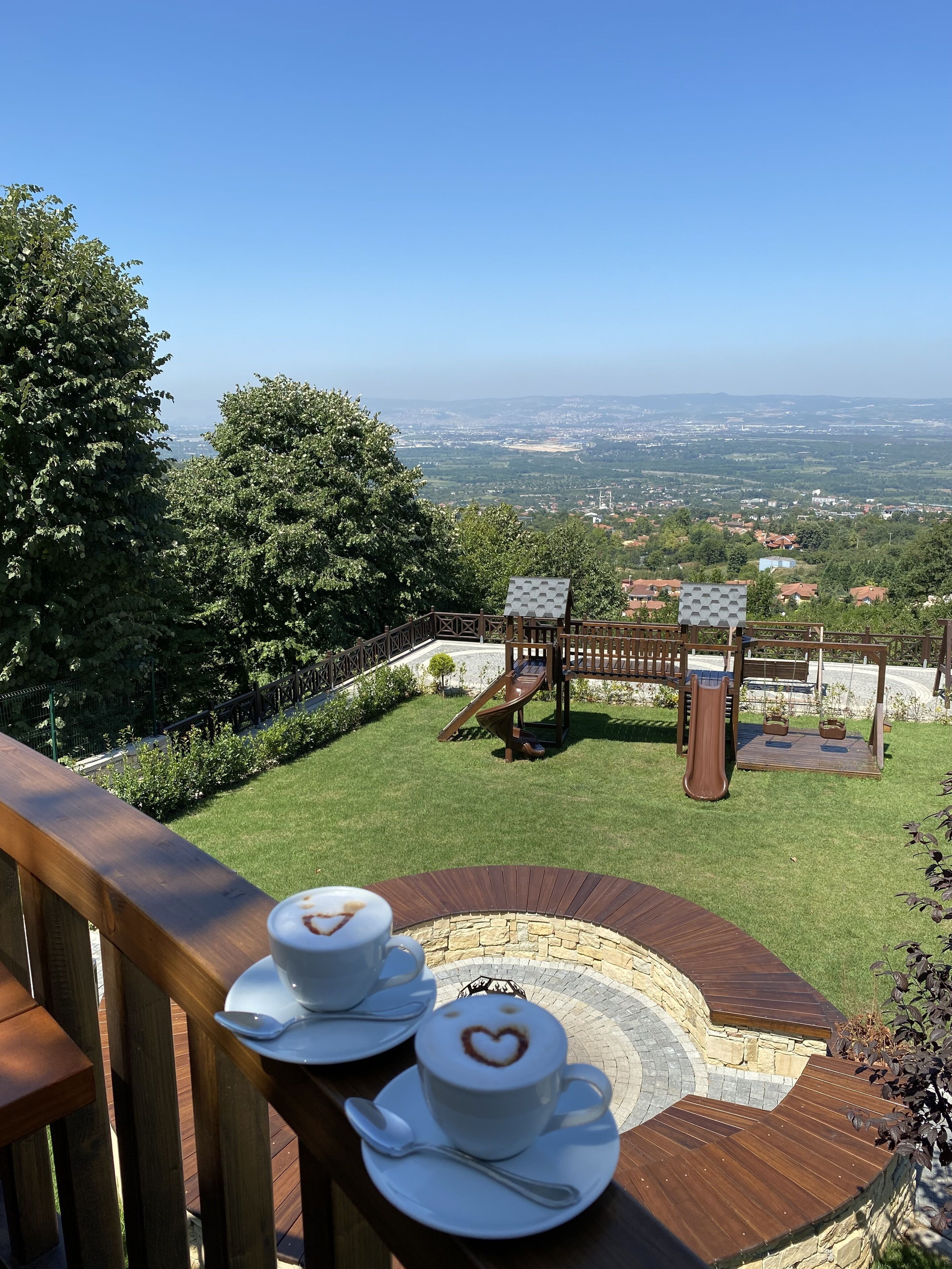 Une terrasse offrant une vue magnifique, deux tasses de café décorées de motifs en forme de cœur, et un terrain de jeux en arrière-plan.