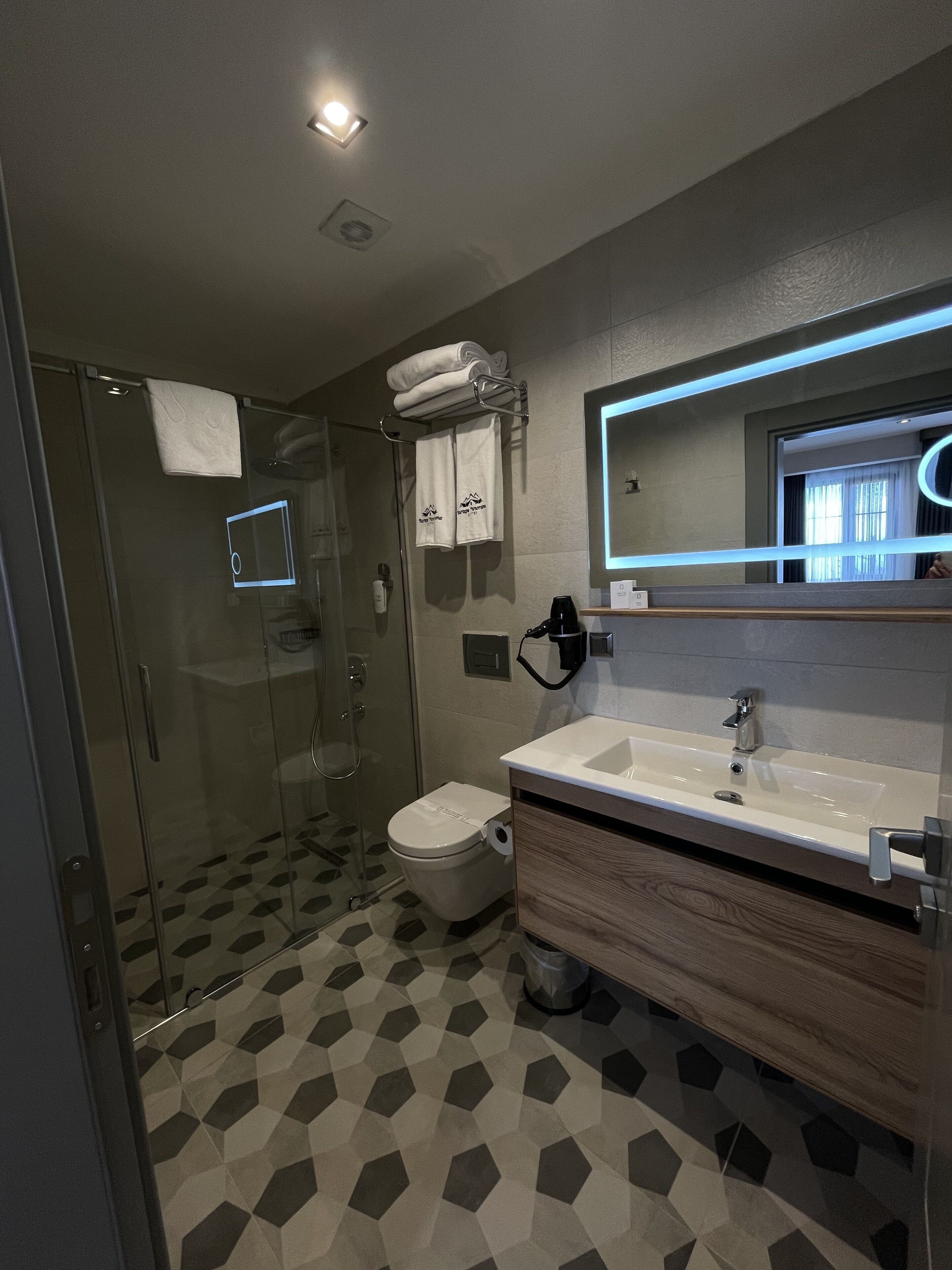 A modern bathroom with a glass shower enclosure, a toilet, a sink with a mirror, and a patterned floor.