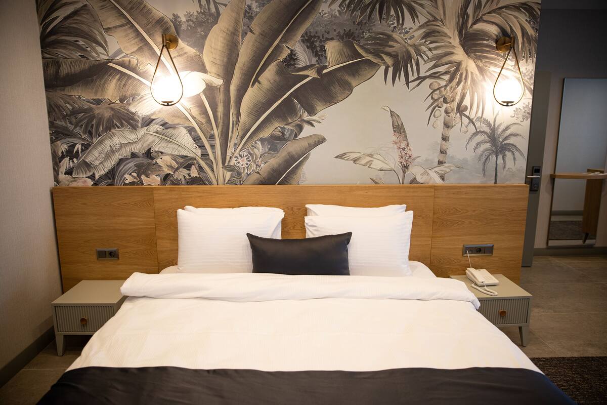 Un lit bien fait, avec une literie blanche et noire, deux lampes de chevet, une table de chevet avec un téléphone et une fresque murale représentant un feuillage tropical.