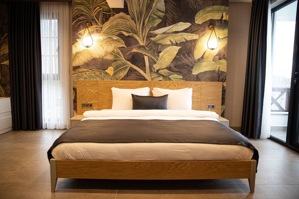 Une chambre d’hôtel moderne avec un grand lit, une tête de lit en bois et une fresque murale représentant des feuilles tropicales.