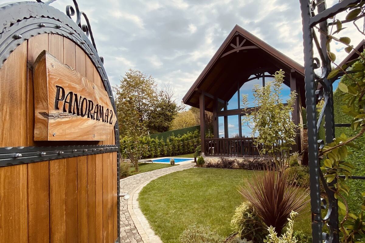 Une porte en bois portant l’inscription « PANORAMA 2 » mène à un bâtiment doté d’un patio couvert et d’une piscine.