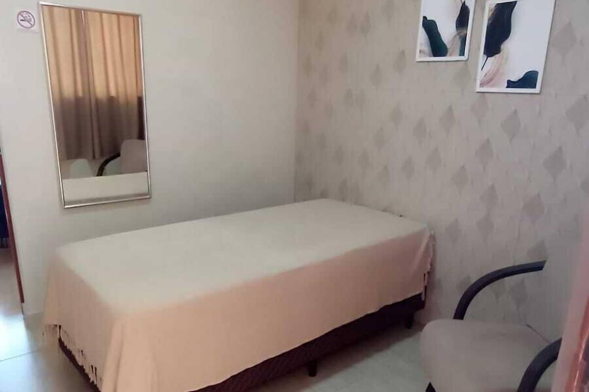 Un dormitorio pequeño y ordenado con una cama individual, una silla, un espejo y dos cuadros colgados en la pared.