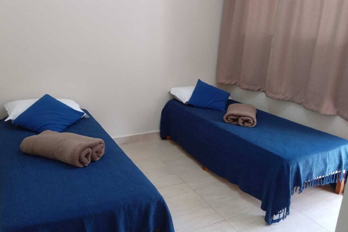 Dos camas individuales con ropa de cama y almohadas azules, un espejo a la izquierda y una ventana con cortinas.