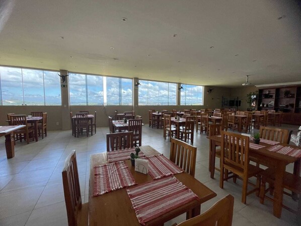 Un comedor amplio con varias mesas y sillas de madera, ventanas grandes que ofrecen vistas al cielo y nubes, y un ventilador de techo.