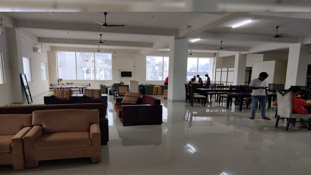 A spacious, well-lit indoor area with high ceilings, ceiling fans, and large windows. There are several seating areas with couches and chairs, and a group of people gathered around a table. - Abuzz Oxfordcaps Dehradun - Campus Accommodation (Dehradun)