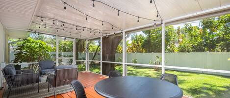 A covered patio with a round table and chairs, surrounded by a wooden deck and a white fence.