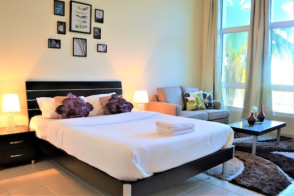 A neatly made bed with white linens, a black headboard, and decorative pillows. There is a nightstand with a lamp, a small table with two vases, and a sofa with cushions. The room has a large window with curtains, and artwork is displayed on the wall.