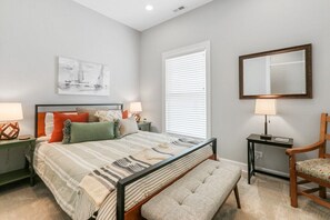 Un dormitorio con cama, mesitas de noche, un banco, una silla, un espejo y una ventana con persianas.