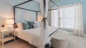 Een hemelbed met een baldakijn, een nachtkastje met een lamp, een decoratief wandobject en een raam met gordijnen.