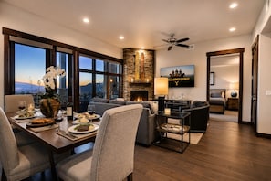 A modern living room with a fireplace, dining area with a table set for a meal, and a large window with a view.