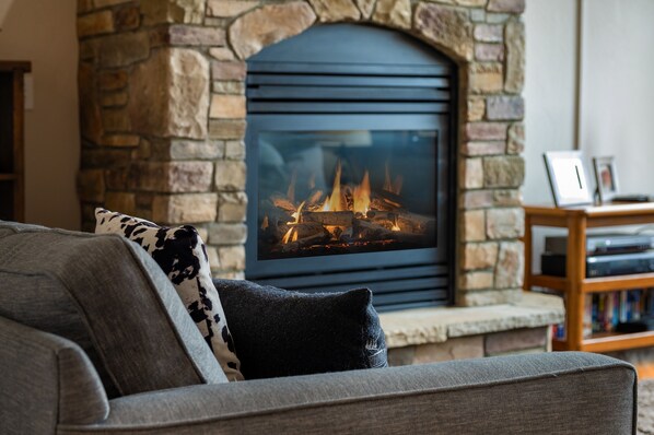 A stone fireplace with a glass front, a grey sofa with patterned cushions, and a wooden bookshelf in a living room.