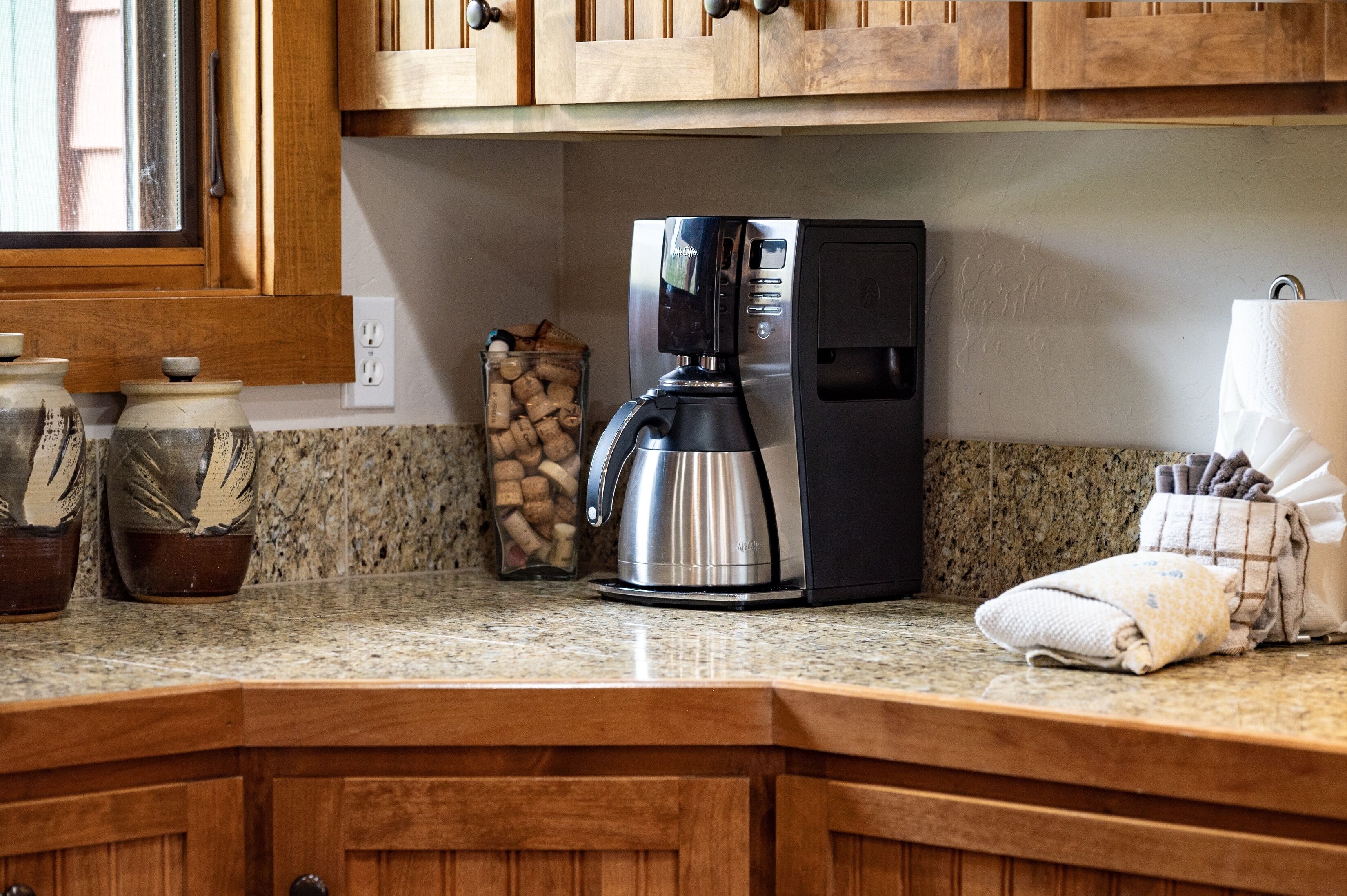 A kitchen with wooden cabinets, a coffee maker, a glass jar of corks, and a towel on the counter.
