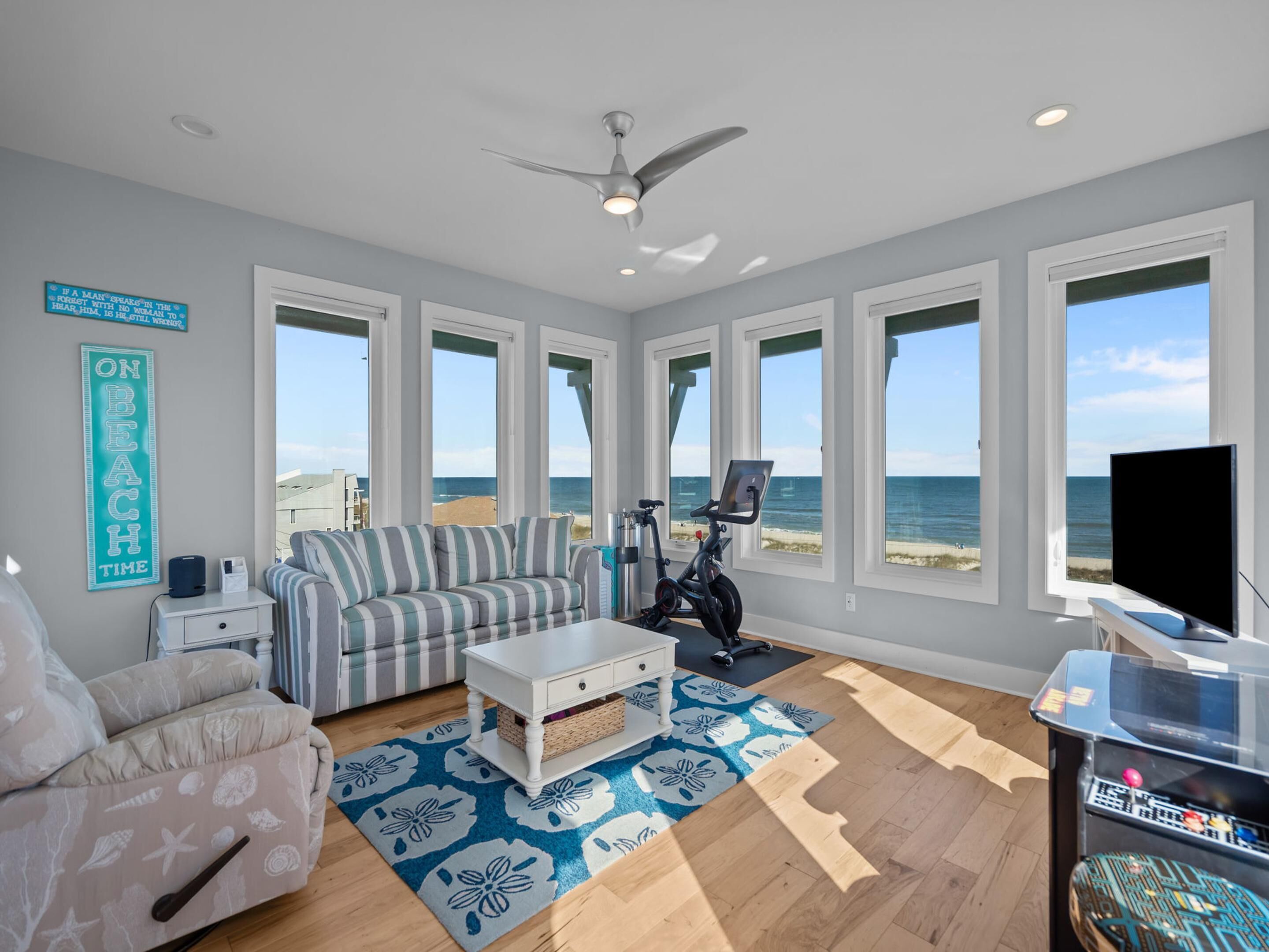 A spacious living room with a large window overlooking the beach, a ceiling fan, and a flat-screen TV.