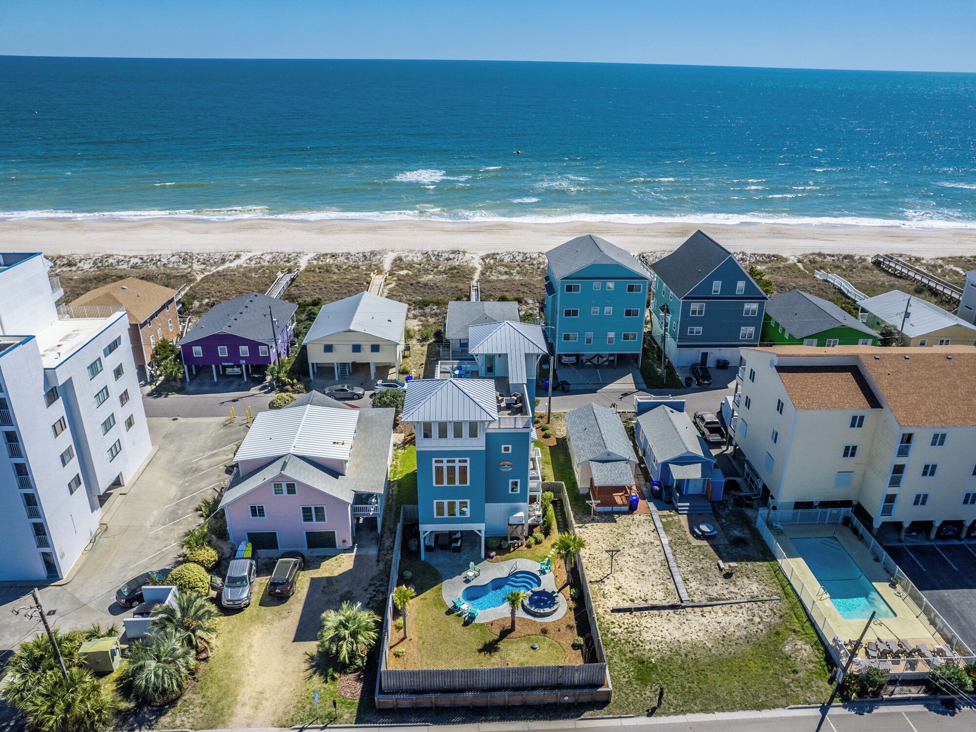 An aerial view of a coastal residential area with houses, a swimming pool, and a beach in the background.