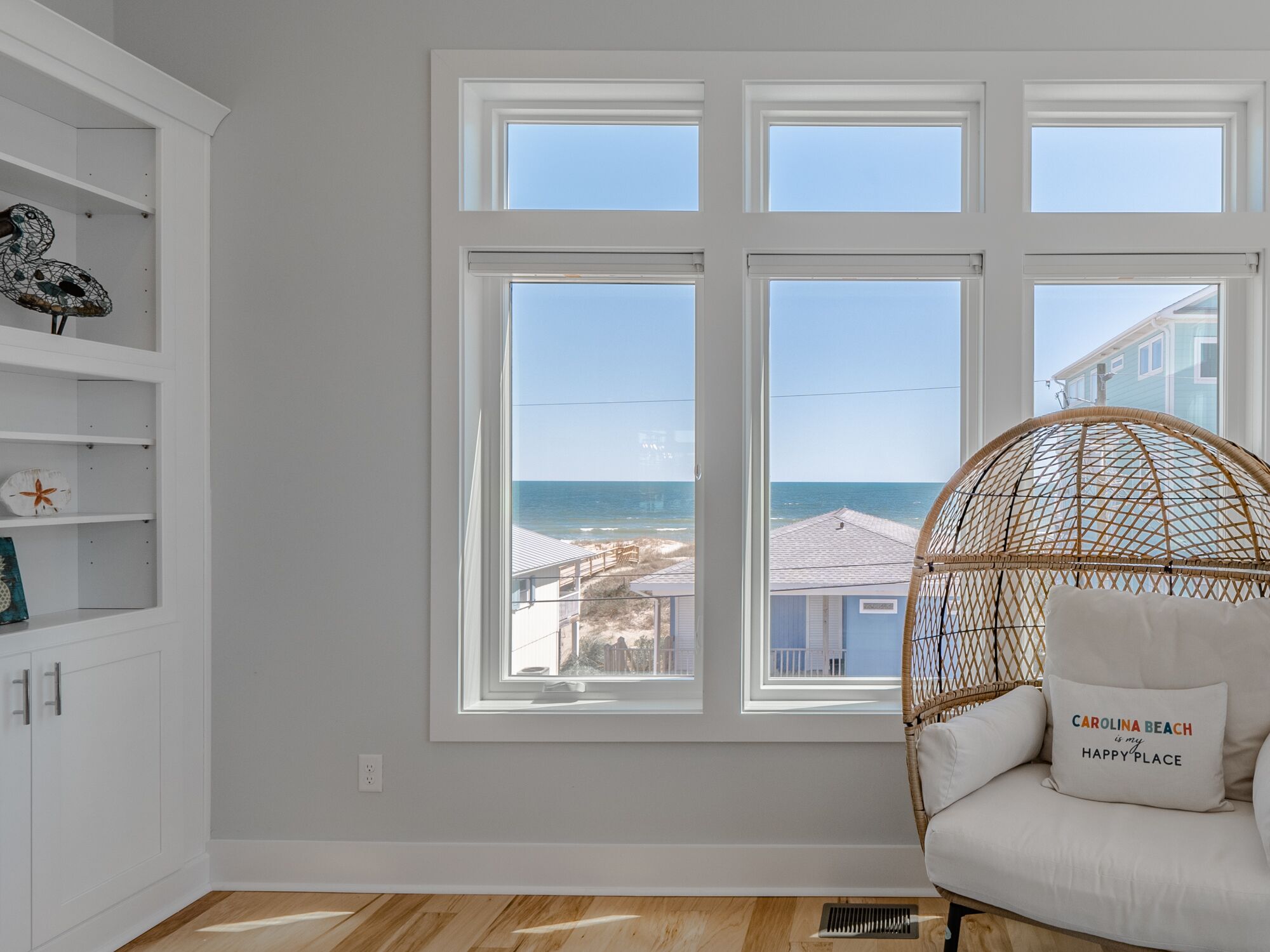 Habitación con un ventanal grande con vistas a la playa, una silla de mimbre con cojín y estantes blancos con objetos decorativos.