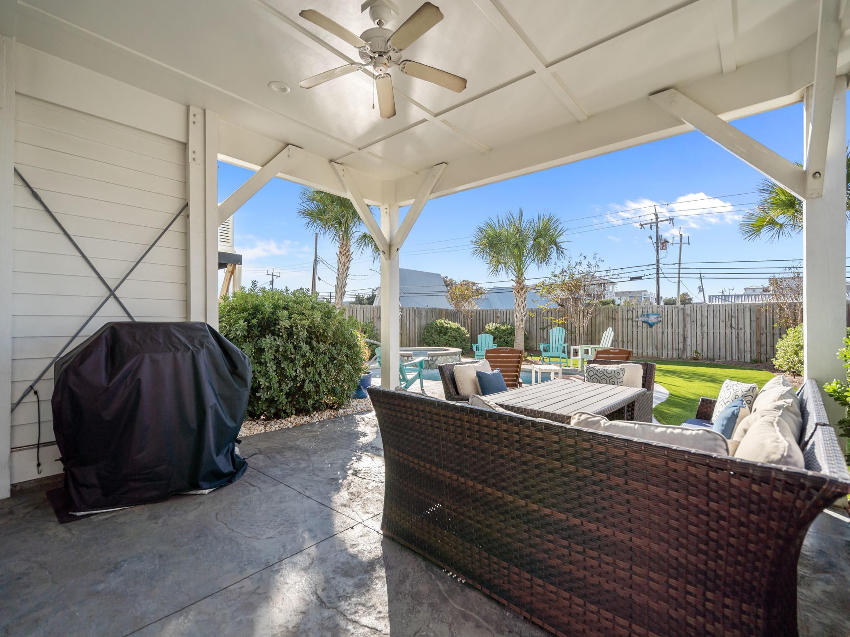 A covered patio with a ceiling fan, outdoor furniture, and a view of a backyard with a pool and palm trees.