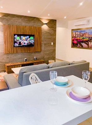Un salon moderne avec un téléviseur à écran plat fixé sur un mur texturé, un ensemble de canapés, une table à manger avec de l’assiette et des verres, et un tableau représentant un paysage urbain.