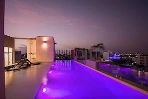 Un espace piscine aménagé sur le toit, avec une longue piscine étroite illuminée de lumières violettes, entourée d’un petit muret et de palmiers.