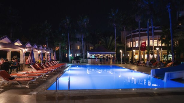 A swimming pool with lounge chairs and umbrellas, surrounded by palm trees and a building with a bar area. - Nergos Side Hotel (Manavgat)
