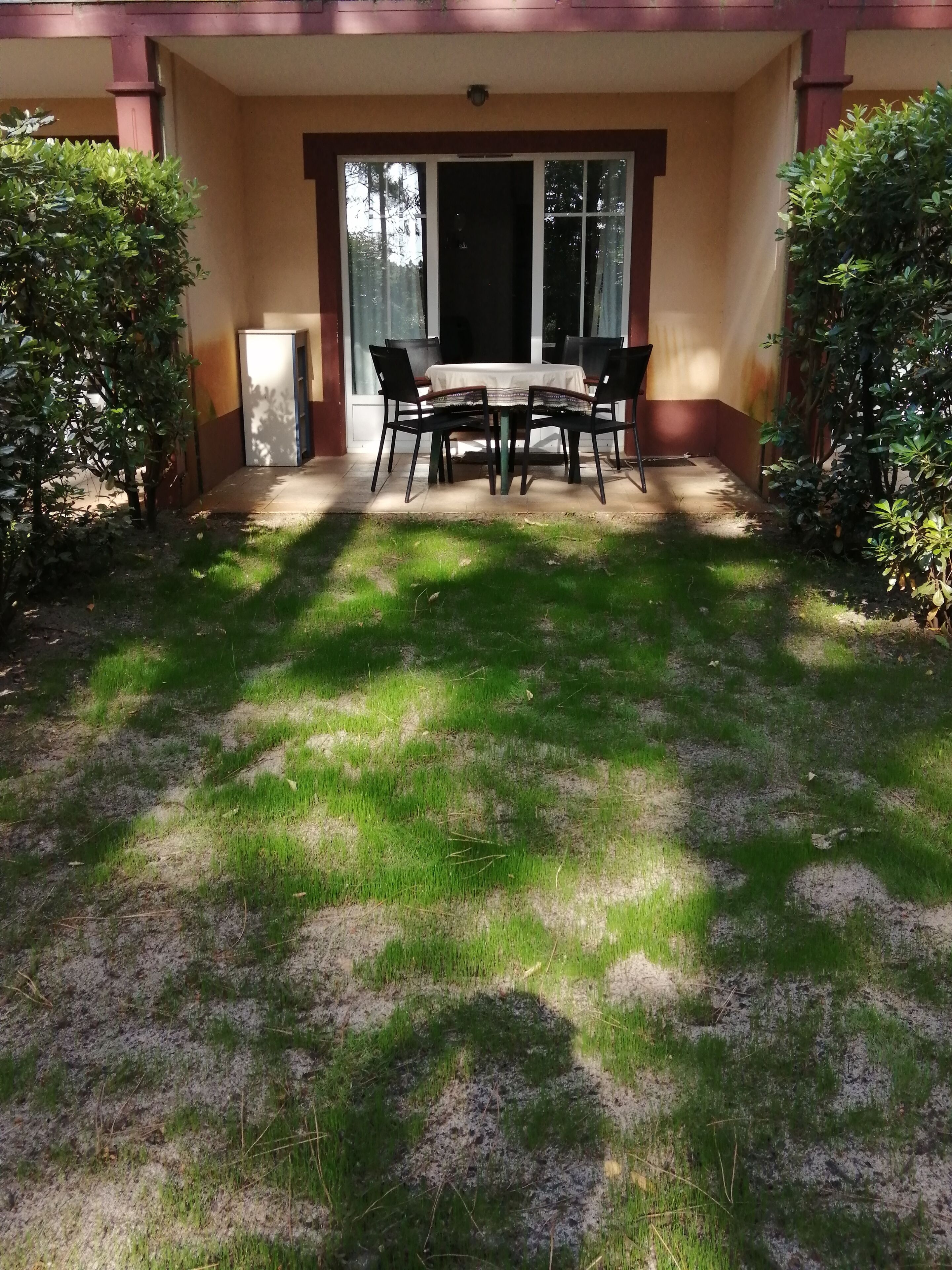 A covered patio with a table and chairs, surrounded by greenery and a door leading to a building.