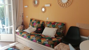 A cozy room with a sofa bed, a small table, and a chair. There are decorative wall mirrors and a window with curtains.
