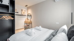 Et moderne hotelværelse med en pænt redt seng, et garderobeskab med ophængt tøj, et sminkebord med spejl og en aircondition-enhed på væggen.