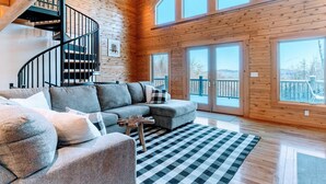 A modern log cabin interior with a curved staircase, a sectional sofa, and large windows offering a view of the outdoors. - Luxury White Mountain Adventure Cabin & Sauna (Bethlehem)