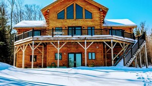 A large wooden cabin with a balcony, a covered porch, and a staircase leading to the upper level. - Luxury White Mountain Adventure Cabin & Sauna (Bethlehem)