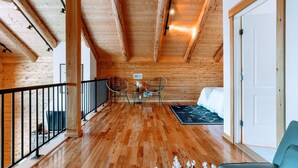 A spacious room with wooden flooring, a balcony with a black railing, two wicker chairs, a small table with a vase, and a bed with a black and white patterned rug. - Luxury White Mountain Adventure Cabin & Sauna (Bethlehem)