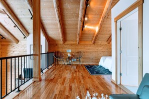 Una habitación amplia con suelo de madera, un balcón con barandilla negra, dos sillas de mimbre, una mesita con un florero y una cama con alfombra de diseño blanco y negro.