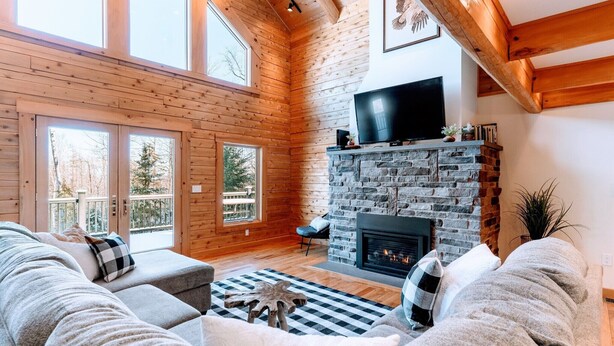 A cozy living room with a stone fireplace, wooden beams, a large TV, and a comfortable seating area. - Luxury White Mountain Adventure Cabin & Sauna (Bethlehem)