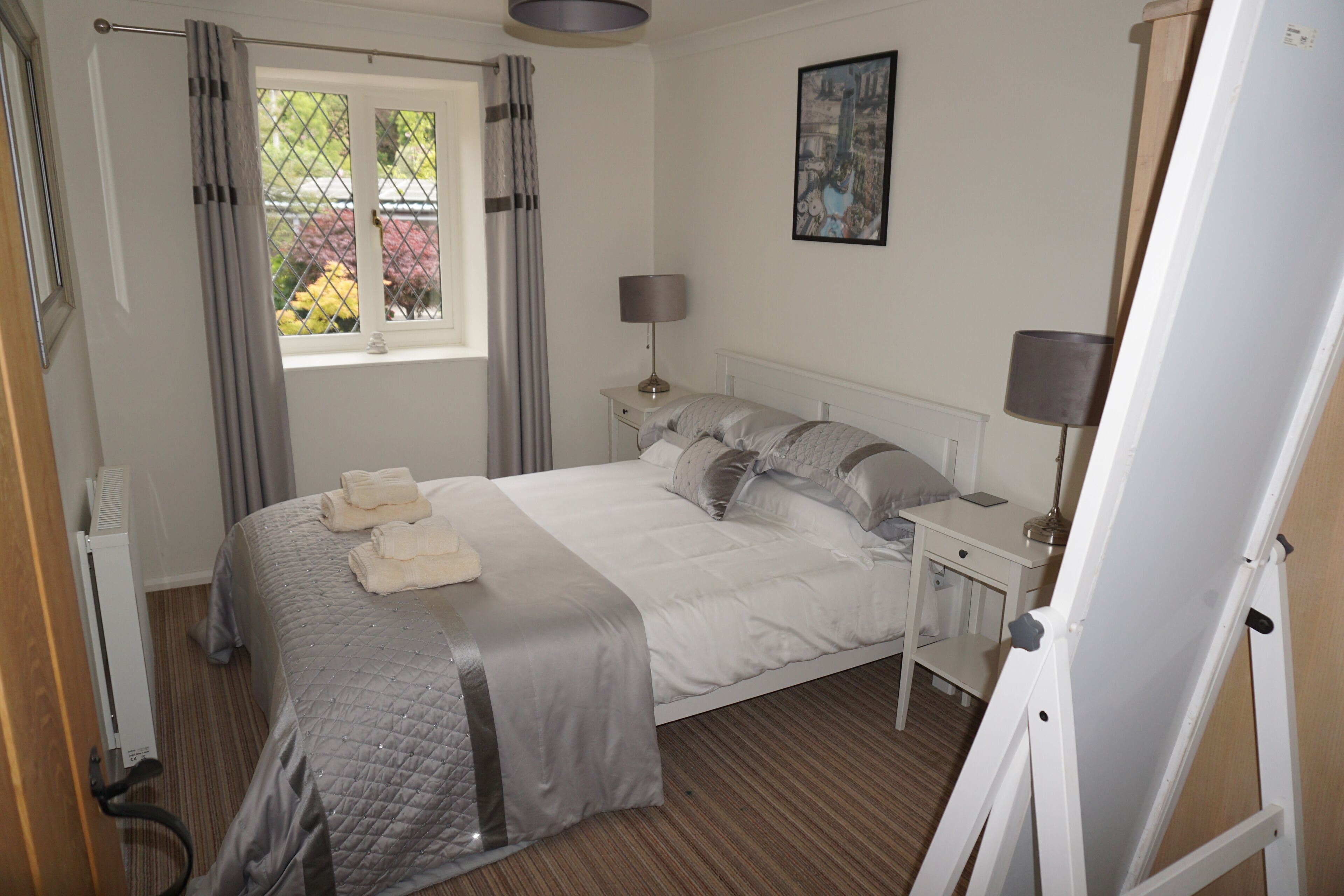 Une chambre bien rangée, avec un lit, des tables de chevet, une fenêtre avec des rideaux, un radiateur et un tableau accroché au mur.