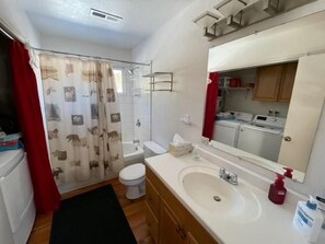 Un baño pequeño con ducha, inodoro, lavamanos y un espejo que refleja una zona de lavandería.