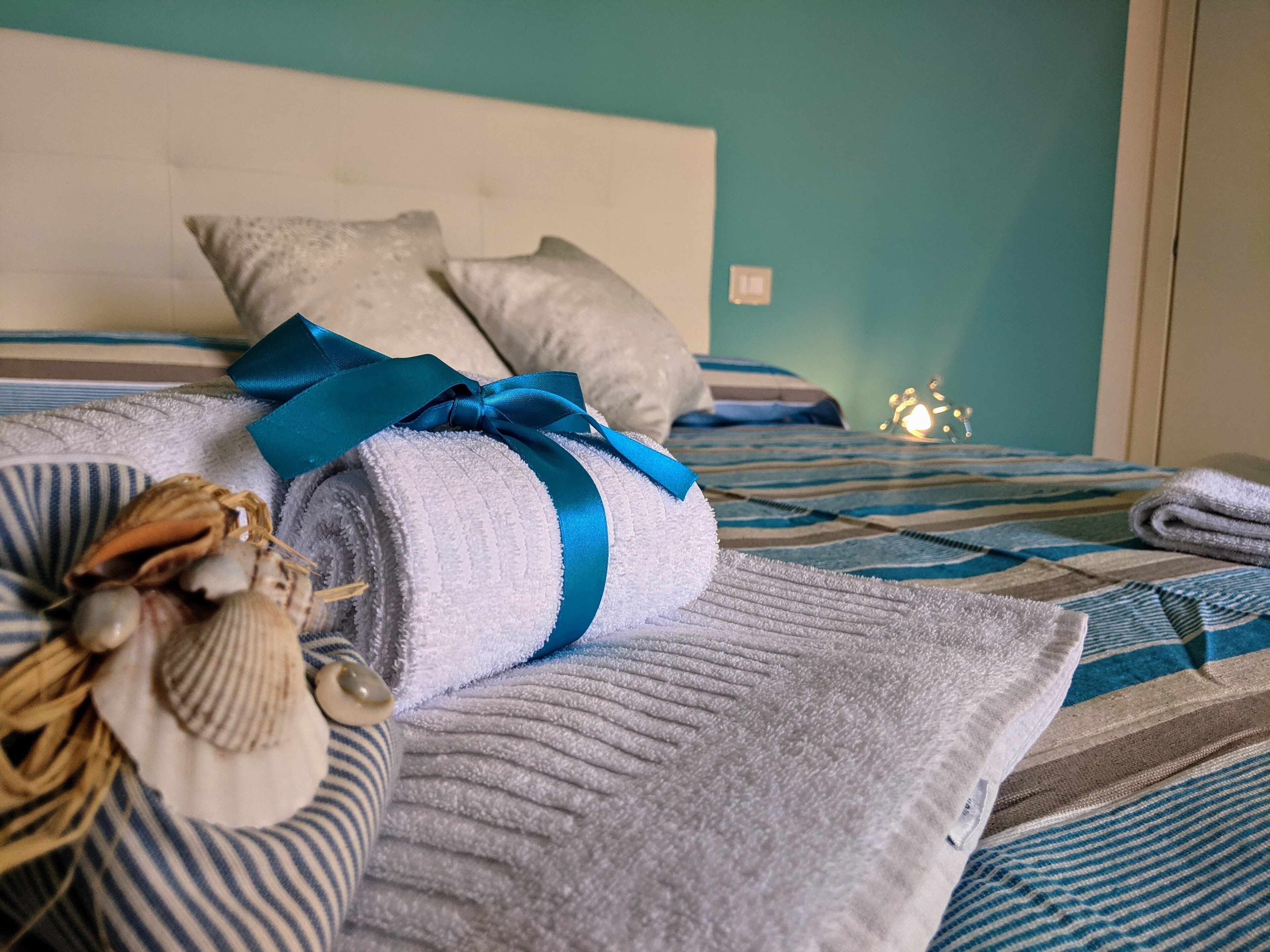 A neatly made bed with a striped blanket, a rolled towel with a blue ribbon, and decorative seashells.