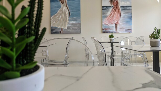 Un comedor con una mesa de mármol y sillas transparentes, dos cuadros de una mujer con vestido y una planta de cactus.