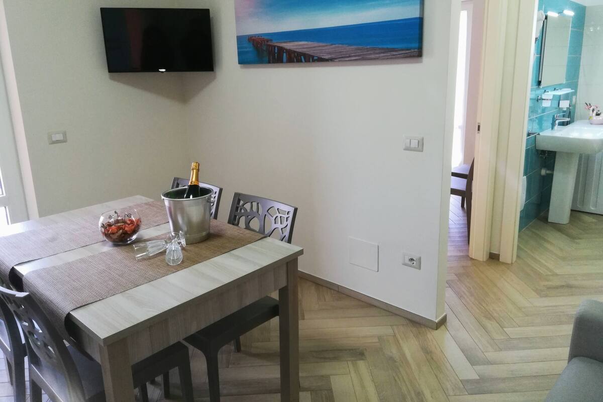 A dining area with a wooden table, chairs, a TV mounted on the wall, a painting of a pier, and a view into a bathroom with a sink and mirror.
