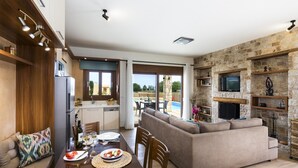 A modern kitchen with a dining area, a stone fireplace, and a view of a swimming pool. - Muses Beach Villas - Erato (Sitia)