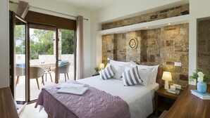 A bedroom with a stone wall, a bed with white bedding and striped pillows, a nightstand with a lamp, and a view of the outdoors through a glass door. - Muses Beach Villas - Erato (Sitia)