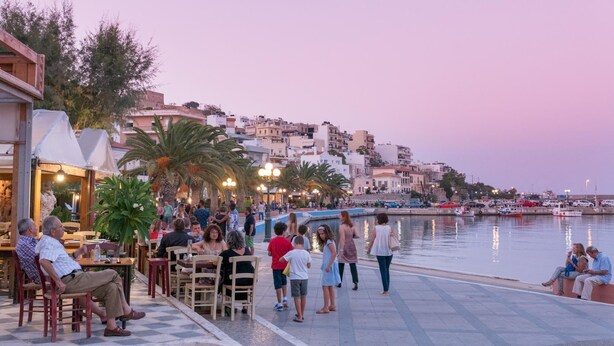 A waterfront promenade with people dining and walking along the path, overlooking a marina and buildings in the background. - Muses Beach Villas - Erato (Sitia)