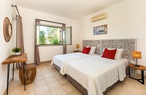 Camera d'albergo con due letti, una finestra che si affaccia sugli alberi, un condizionatore d'aria a parete e un comodino con una lampada.