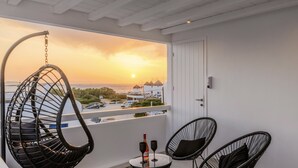 A balcony with a hanging chair, two chairs, a table with a bottle and glasses, and a view of the sunset over the sea. - Sunset Windmills Suites (Mykonos)