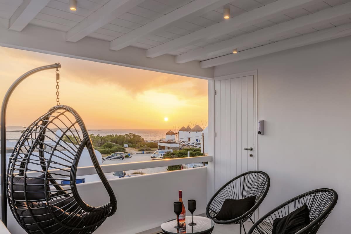 A balcony with a hanging chair, two chairs, a table with a bottle and glasses, and a view of the sunset over the sea.