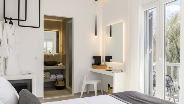 A modern bedroom with a large bed, a small desk, a mirror, and a view of the outdoors. - Sunset Windmills Suites (Mykonos)