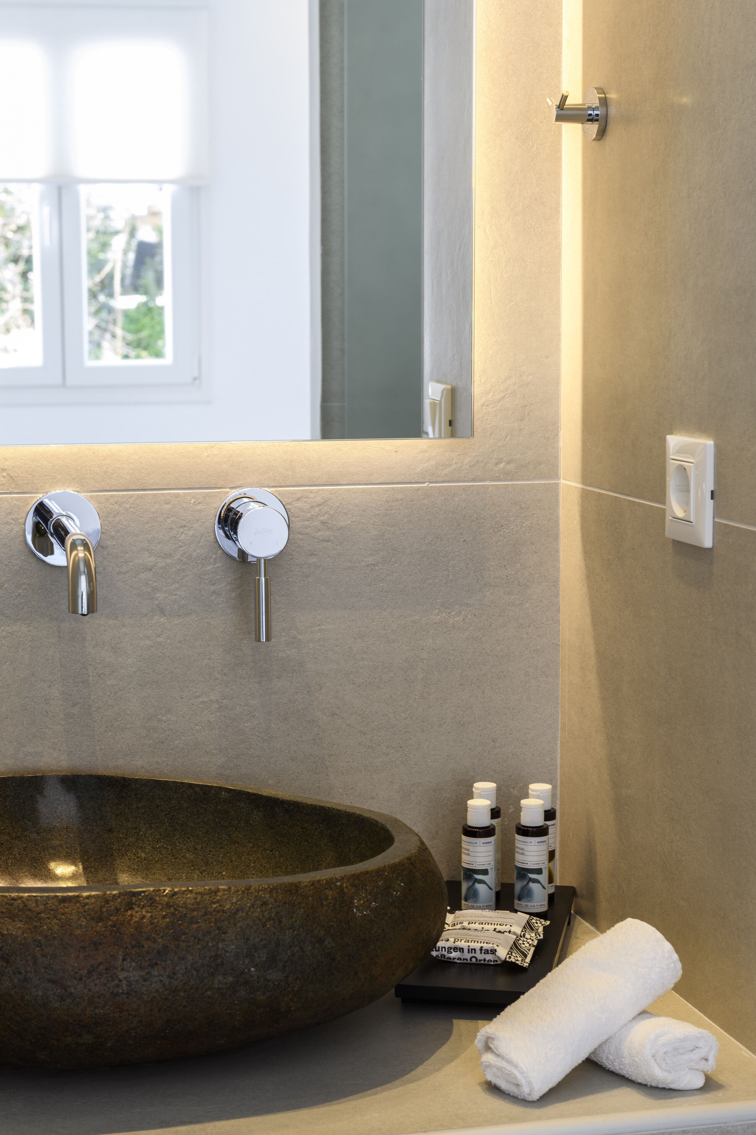 Un baño con un lavabo grande y redondo, dos grifos cromados y un espejo sobre el lavabo. Hay dos botellas de loción y una toalla blanca doblada en la encimera.