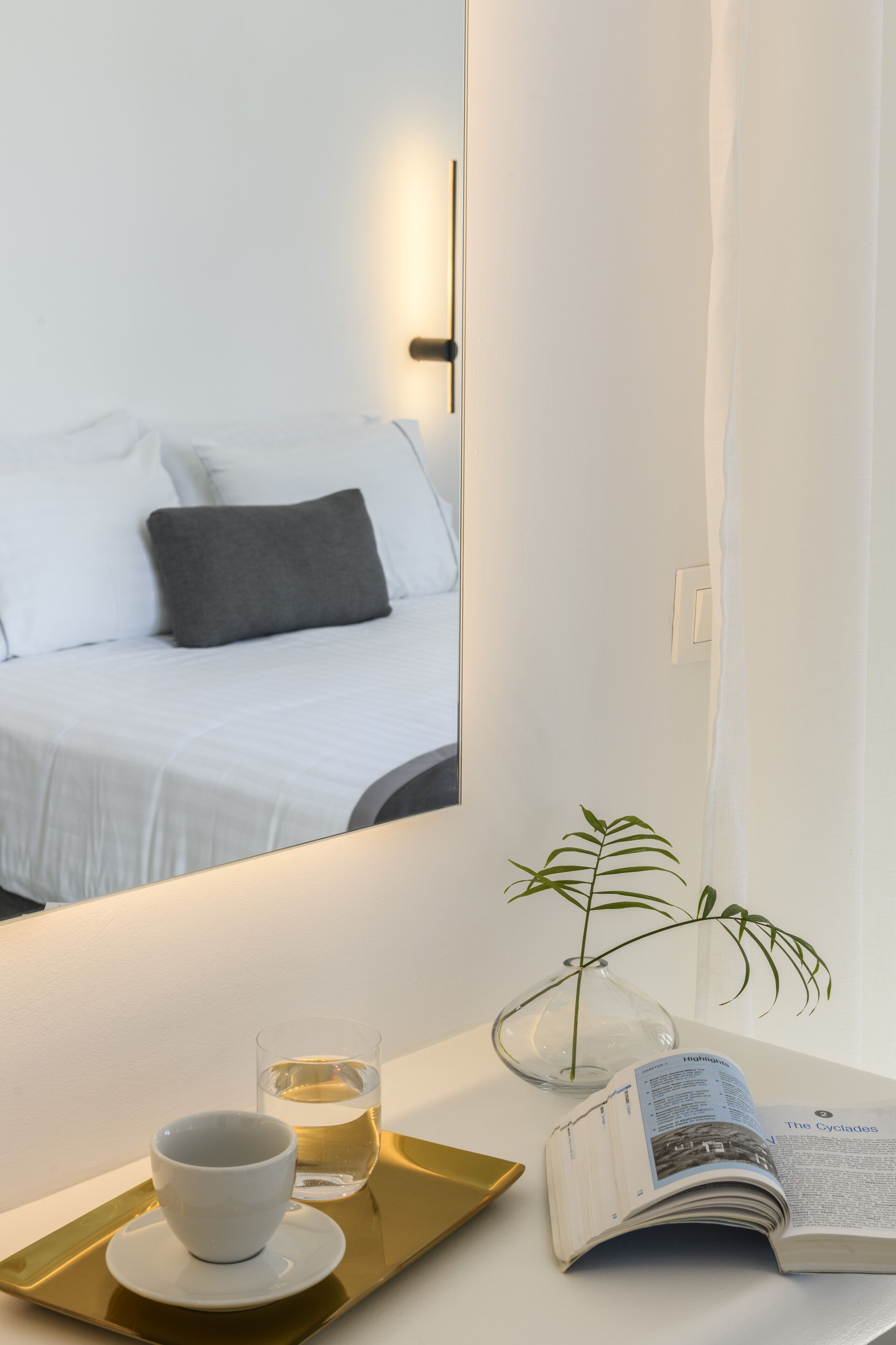 A hotel room with a bed, a bedside table with a glass of water, a cup, a tray, a book, and a potted plant.