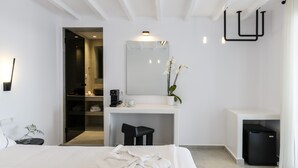 A modern hotel room with a bed, a desk, a mirror, a small stool, a trash bin, and a potted plant. - Sunset Windmills Suites (Mykonos)