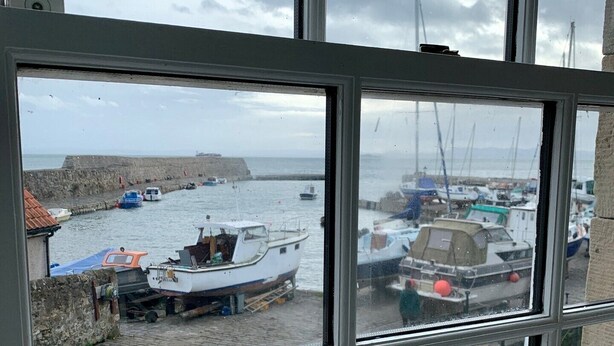 Blick durch ein Fenster auf einen Hafen mit vor Anker liegenden Booten, eine Steinmauer und ein bewölkter Himmel.