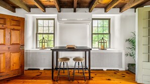 Una stanza con pavimento in legno, un tavolo con due sgabelli, un termosifone e due finestre con stipiti in legno.