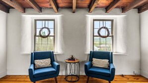 Una stanza con due poltrone blu, un tavolino rotondo con libri e un oggetto decorativo, e due finestre adornate da ghirlande.