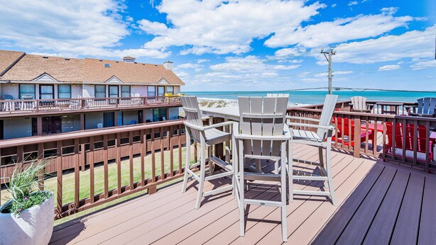 A deck with chairs and a table overlooking a beach, with a building in the background. - OCTIPI Your SenSEAS - Kitty Hawk, Sea Dunes, Open All Year, Flex Stays, Pool, Clubhouse (Kitty Hawk)